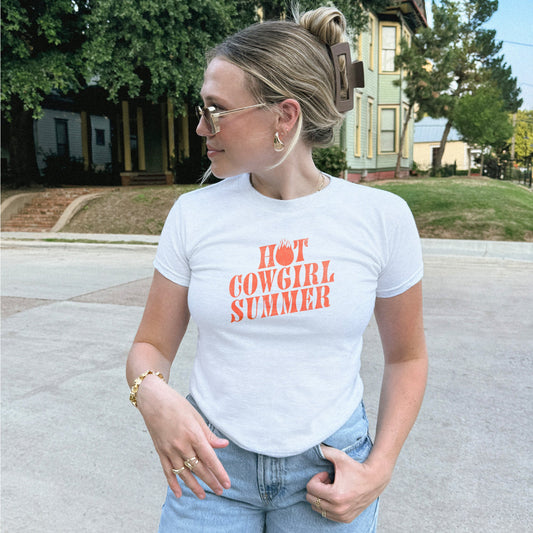 Woman wearing a white t-shirt with 'Hot Cowgirl Summer' text outdoors.