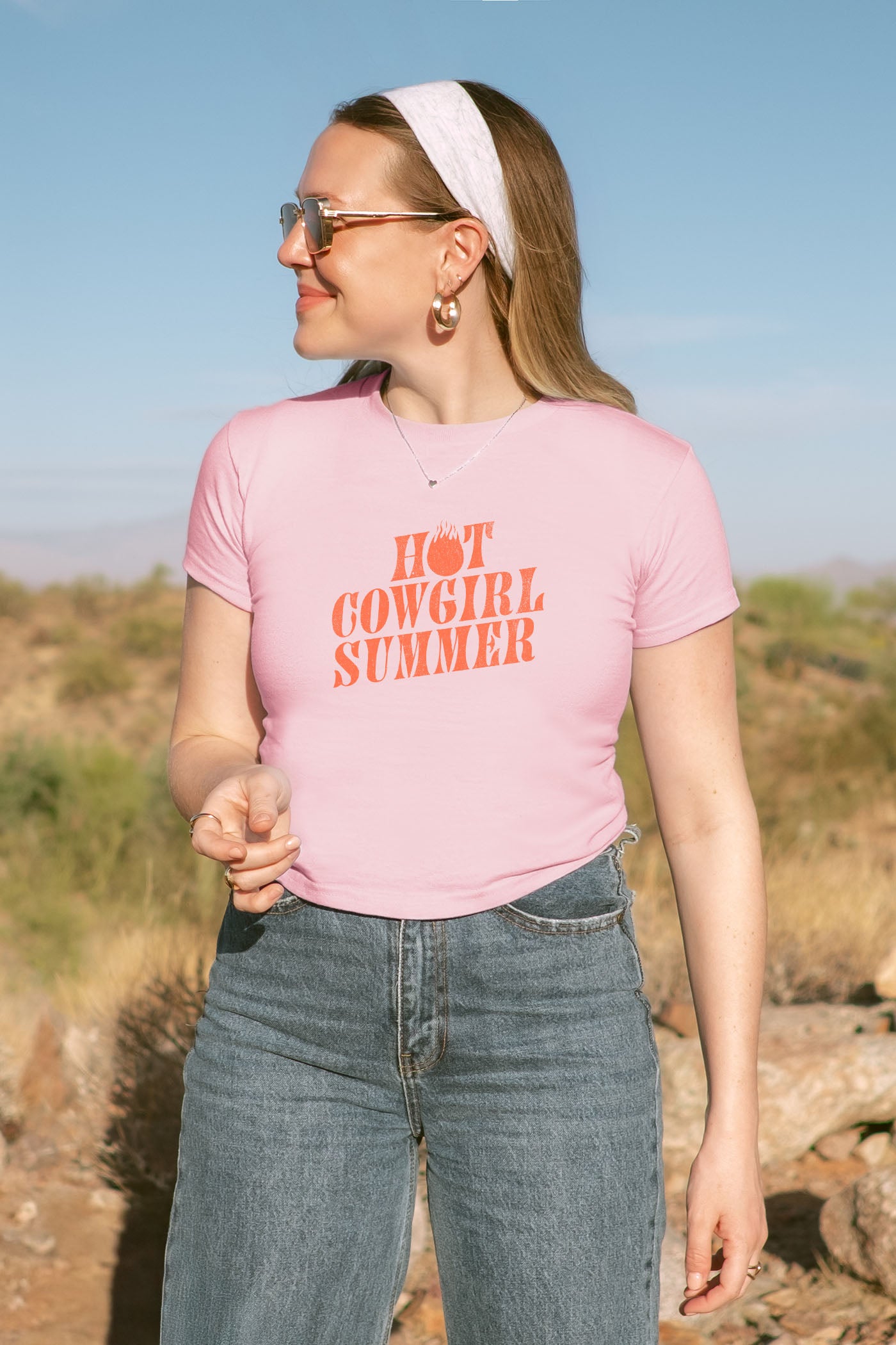 Woman wearing a pink t-shirt with 'Hot Cowgirl Summer' text, standing outdoors.