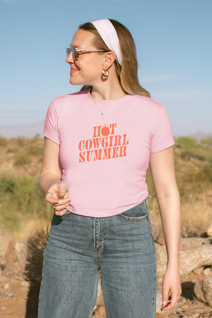 Woman wearing a pink t-shirt with 'Hot Cowgirl Summer' text, standing outdoors.