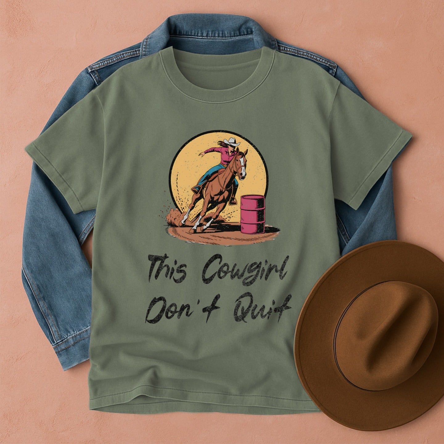 Green t-shirt with a barrel racer cowgirl graphic and text, placed on a denim jacket and brown hat on a peach background.