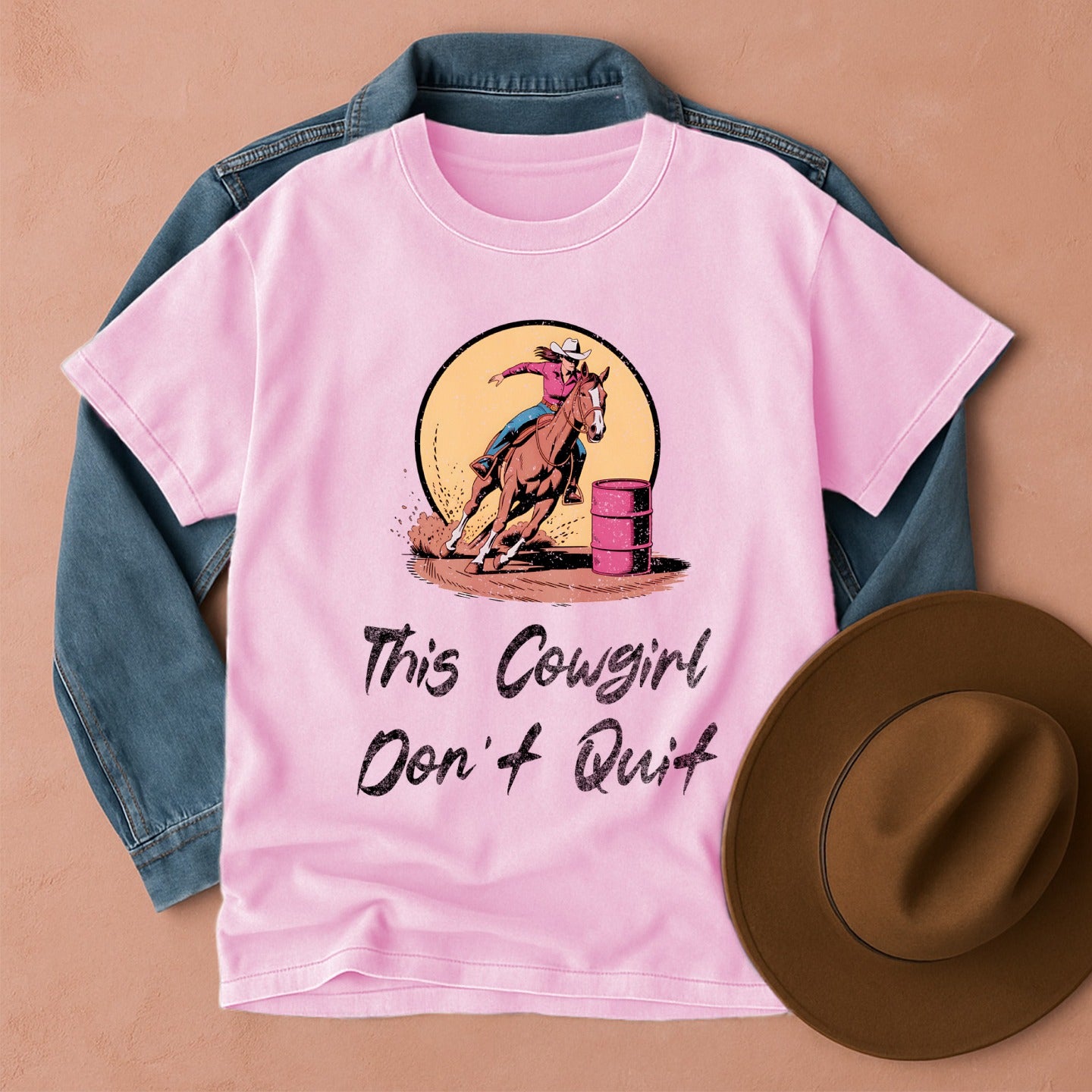 Pink t-shirt with a Barrel Racing Cowgirl  graphic and 'This Cowgirl Don't Quit' text, placed on a denim jacket with a brown hat beside it.