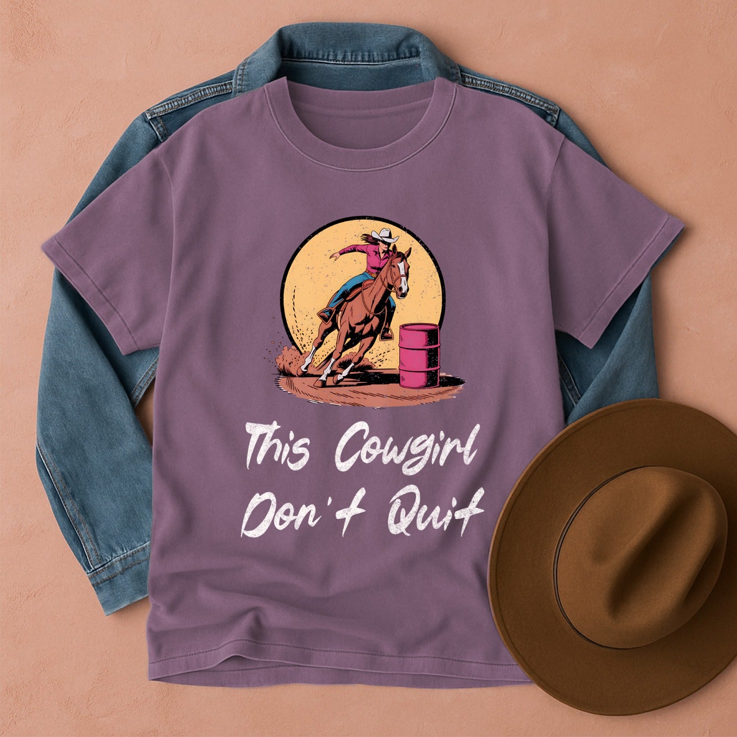 Purple t-shirt with a barrel racer cowgirl graphic and text, placed on denim fabric with a brown hat beside it.
