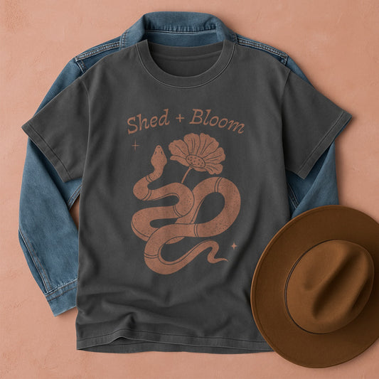 Pepper Comfort Colors t-shirt with snake design and 'Shed + Bloom' text, placed on a denim jacket with a brown hat beside it.
