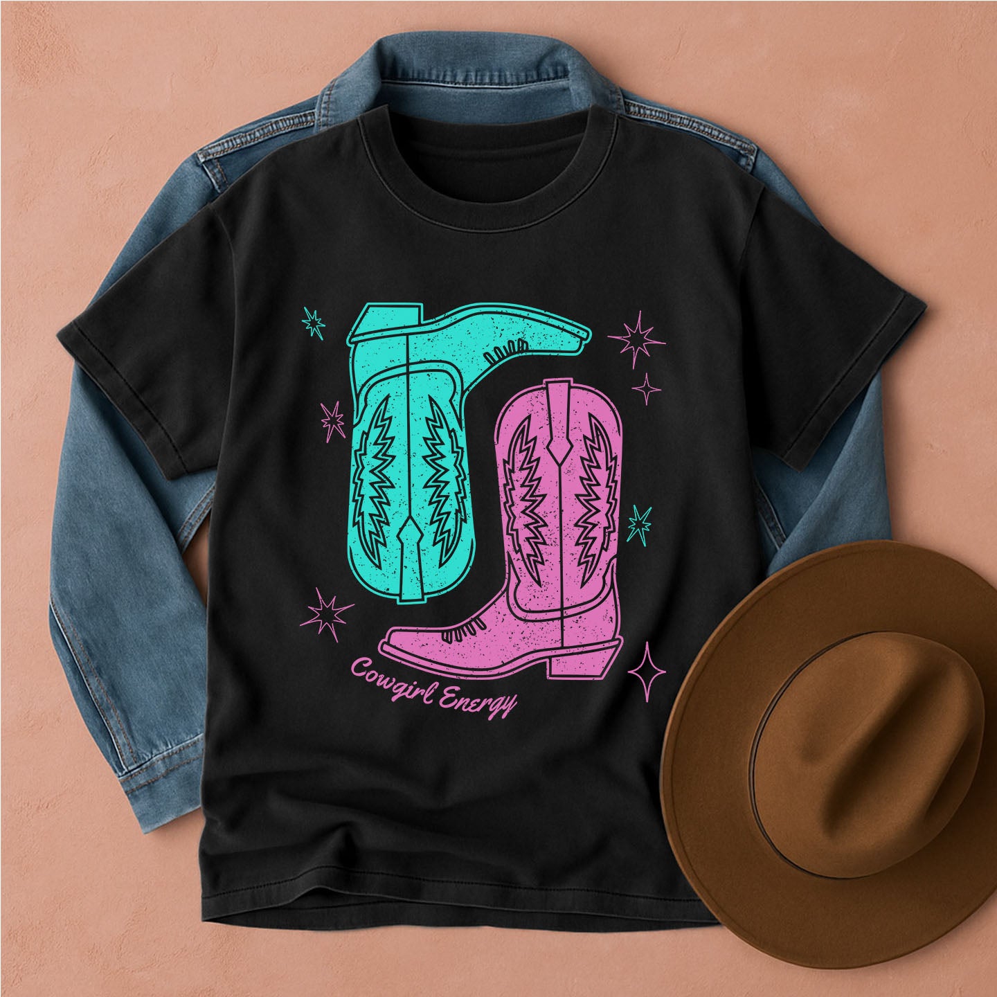 Black t-shirt with cowboy boot design and 'Cowgirl Energy' text, placed on a denim jacket with a brown hat beside it.