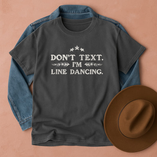 Gray t-shirt with Don't Text. I'm Line Dancing text, blue denim jacket, and brown hat on a beige background