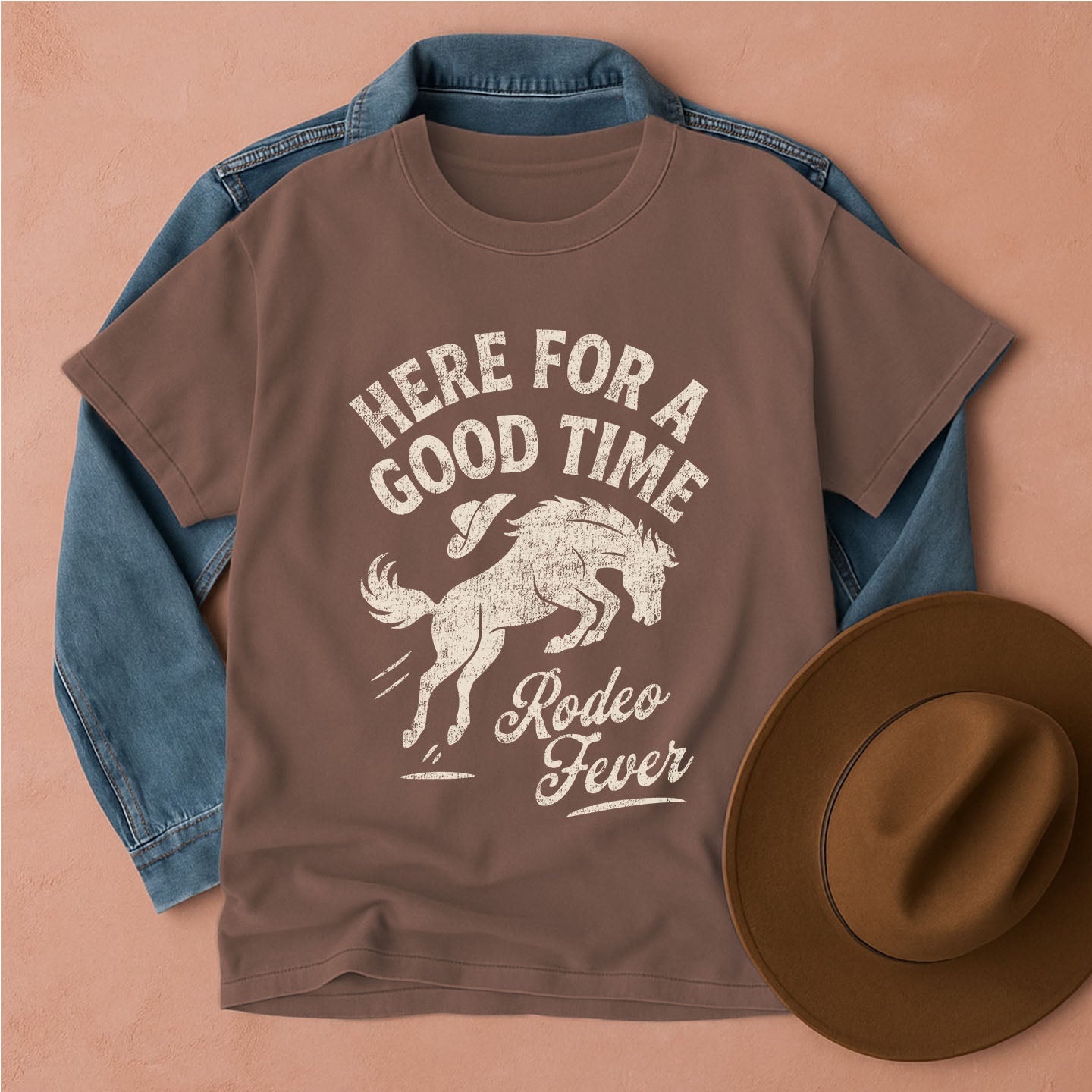 Brown rodeo t-shirt with horse graphic and text, placed on a blue denim jacket and brown hat on a beige background