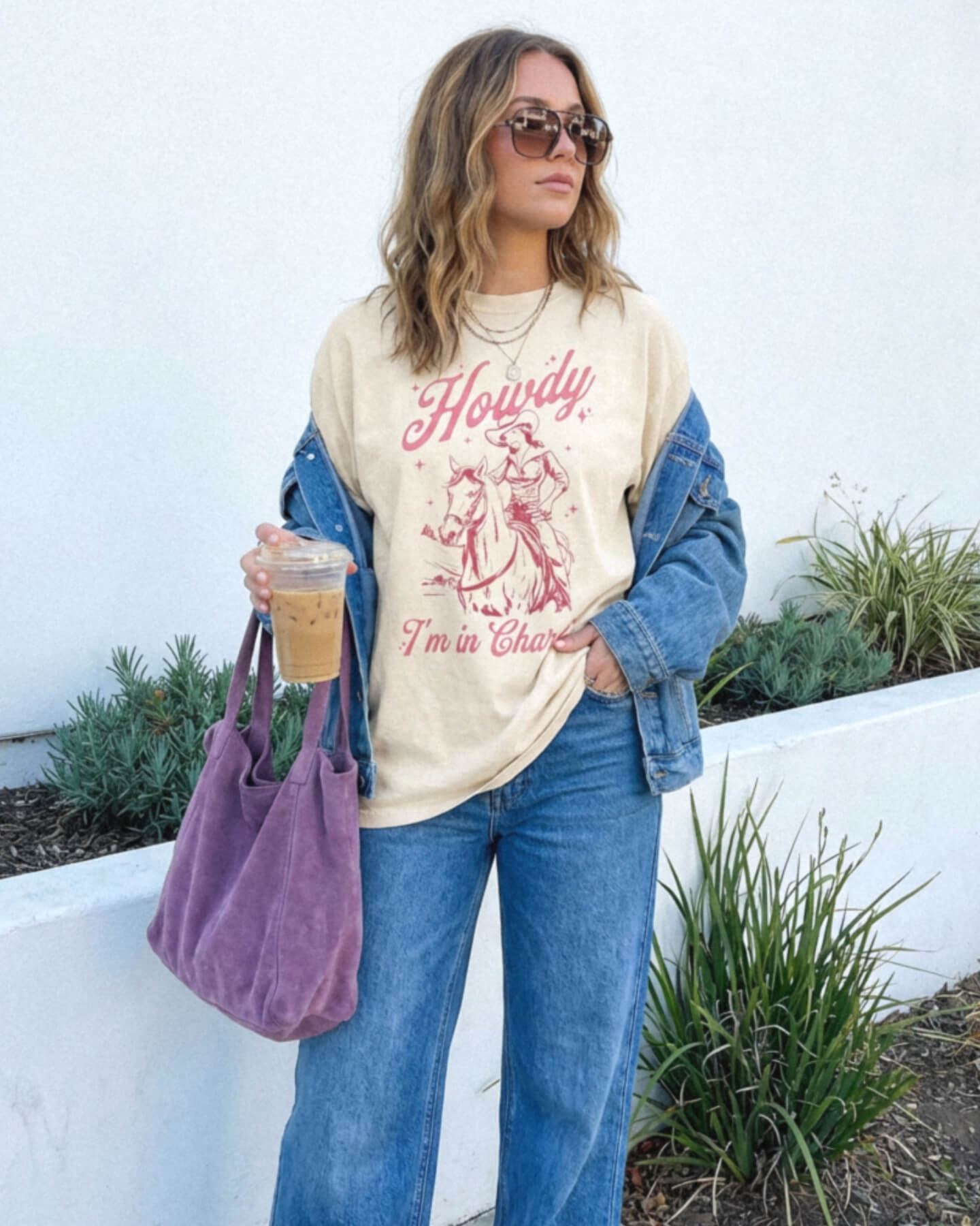 Woman wearing a 'Howdy I'm in Charge' shirt with a horse graphic, holding a purple bag and a drink.