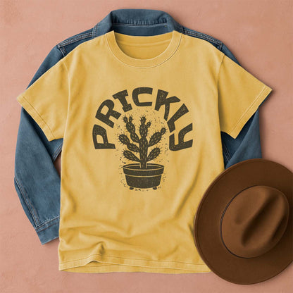 Yellow t-shirt with 'PRICKLY' cactus design, blue denim jacket, and brown hat on a pink background