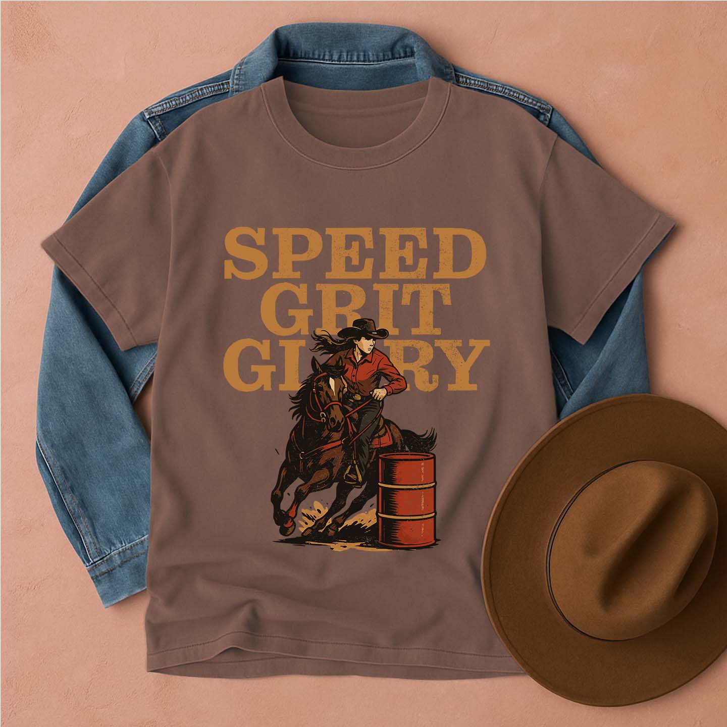 Brown t-shirt with barrel racer cowgirl graphic and 'SPEED GRIT GLORY' text, placed on a peach background with a denim jacket and brown hat.